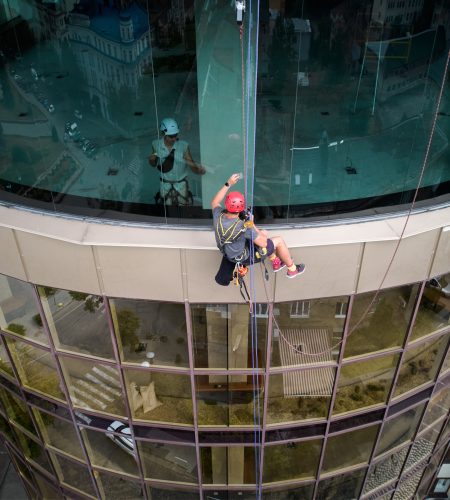Industrial mountaineering worker cleaning window of high-rise building while colleague standing behind glass. Man cleaner using safety equipment while washing skyscraper facade.