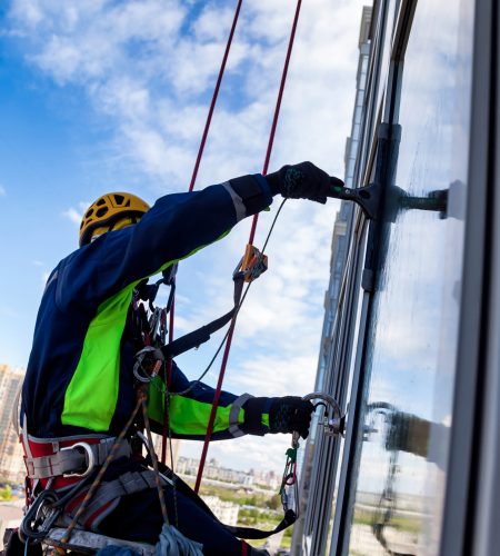 Occupational industrial mountaineering worker washing exterior facade glazing at blue sky backdrop, hanging over building. Rope access laborer climbing on wall of skyscraper. Copy text space for ad